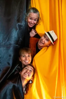 Quatre femmes souriantes pointent leur nez de derrière un rideau jaune