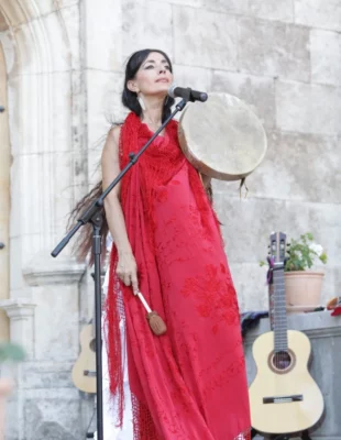 Femme habillée en rouge, débout avec un tambour en main et une guitare derrière elle. Sur le fond une construction ancienne
