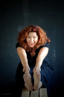 Catherine Lecoq,comédienne,sur un fond noir, assise à jambes allongées, avec les mains qui touchent les pointes des pieds 