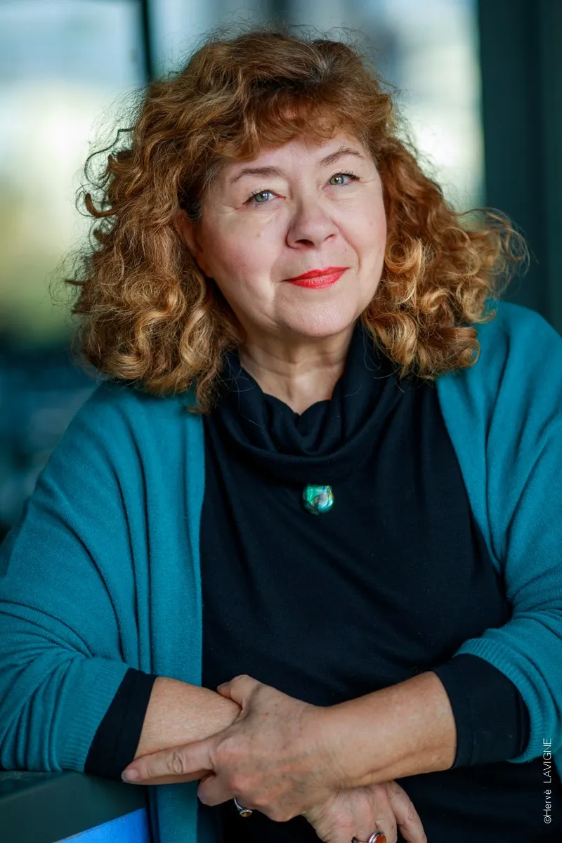 Une femme aux cheveux longs, Catherine Lecoq, habillée en bleu regarde droit dans l'appareil photographique, derrière elle le fond flou d'un extérieur