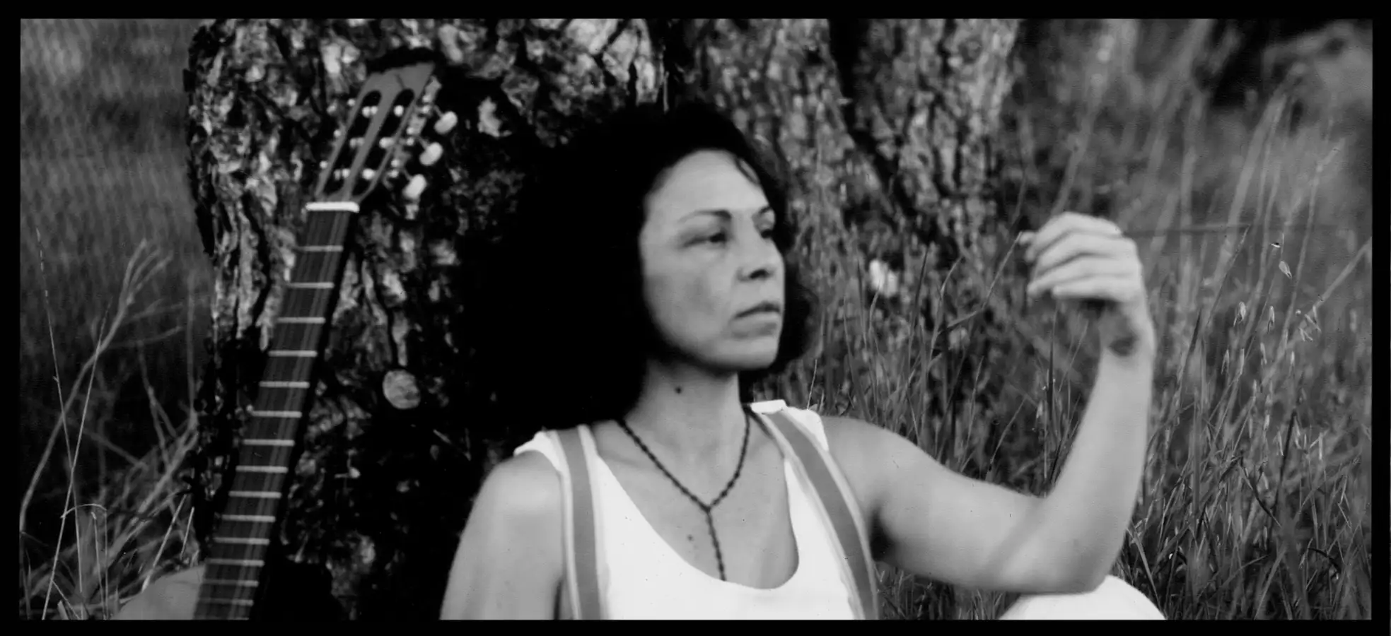 Une femme avec un débardeur blanc est assise le dos contre un arbre à côte d'une guitare