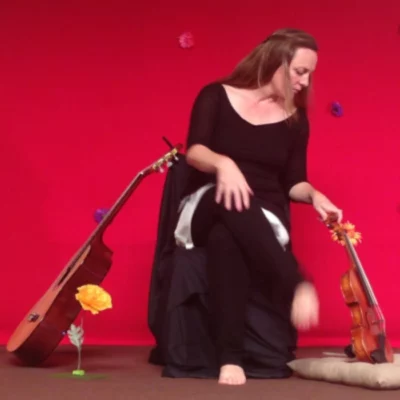 La musicienne Stéphanie Joire assise sur une chaise, sur un fond rouge avec une guitare à sa droite et un violon qu'elle est en train de regarder à sa gauche