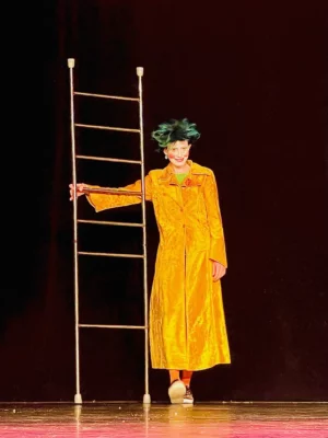 Sur une scène au fond noir, une clown tient une échelle en souriant