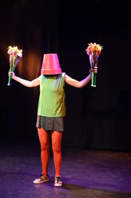 Une personne en short sur une scène de théâtre avec un pot en tête et un bouquet de fleurs par main