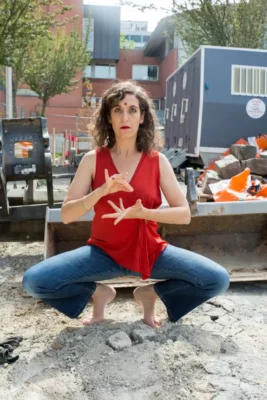 Ofra Hoffman, danseuse et comédienne,en extérieur, dans un paysage urbain : elle est accroupie, avec les bras en position de danse et un cercle rouge au milieu des yeux