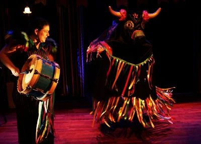 Une femme habillée en costume traditionnel joue un tambour et, à côté d'elle, un personnage en costume de boeuf