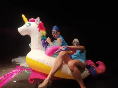 Sur un scène de théâtre, deux femmes en maillot et bonnet de bain, lunettes de natation, sont assises sur un hippocampe en plastique 