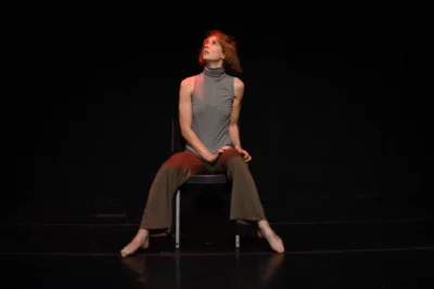 Sur une scène noire, une jeune femme assise sur une chaise regarde vers le haut 