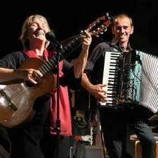 Une femme est en train de jouer la guitare et chanter devant un micro, à ses côtés un homme l'accompagne avec un accordéon