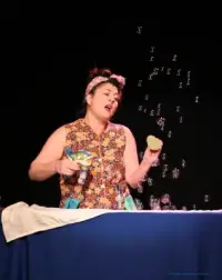 Sur un fond noir, une femme assise à une table bleue est en train de jouer avec des bulles de savon