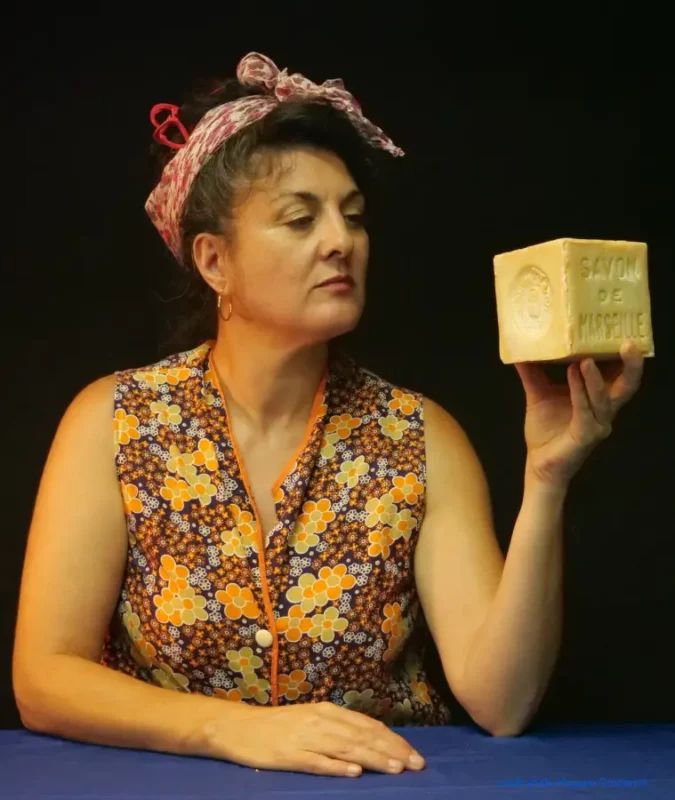 Une femme assise à une table bleu en train de regarder le cube de savon de Marseille qu'elle tient dans sa main