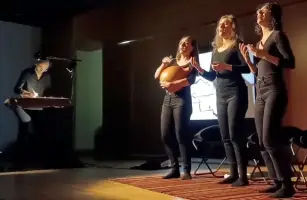 Trois femmes sur scène en train de chanter, un homme penché sur une table de dessin à leur côté