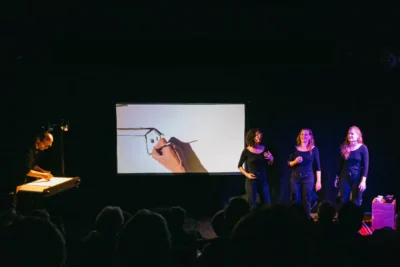 Sur un scène de théâtre, un homme en train de projeter ses dessins sur un écran et trois femmes habillées en noir en train de chanter