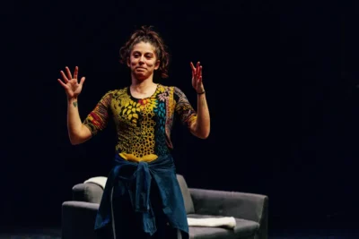 Une jeune femme sur une scène de théâtre au fond noir, elle a les bras ouvert et regarde devant elle en souriant