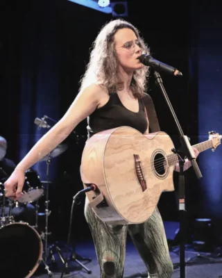 Une jeune femme avec des lunettes de vue, en débardeur et jeans en train de chanter et jouer la guitare devant un micro