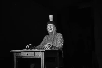 Photo en noir et blanc d'un personnage clownesque assis à une table, derrière un fond noir