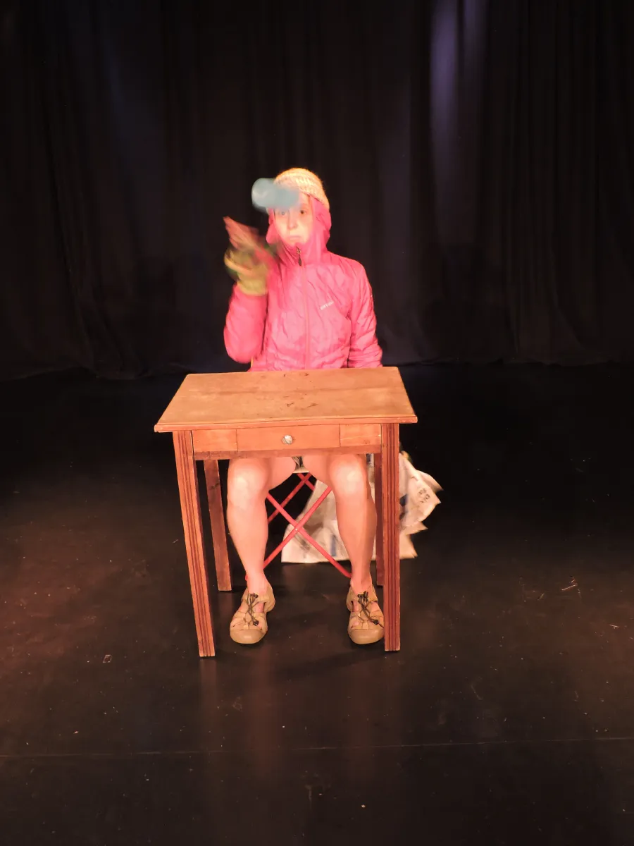 Un personnage clownesque assis à une table, habillé en rose, en train de jongler avec un verre