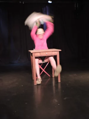 Un personnage clownesque sur une scène de théâtre, derrière une table, les bras en l'air