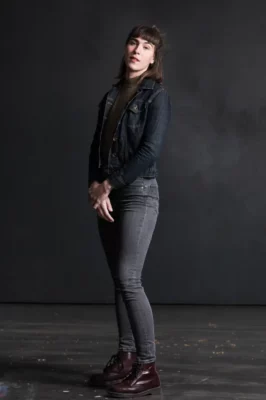 Sur fond noir, une jeune femme de trois quarts, habillée en noir et avec les mains croisée devant elle