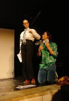 Deux personnes sur une scène de théâtre : un homme à genoux avec des billets dans les mains et une femme, habillé en pantalon et cravate, debout, à ses côtés