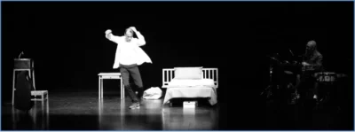 Un homme en chemise blanche en train de danser sur un plateau de théâtre entre en lit et une table