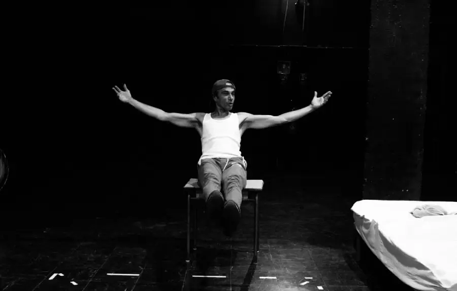 Photo en noir et blanc d'un homme sur un plateau de théâtre. Il est assis sur une table, les jambes allongés devant lui et les bras ouverts