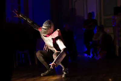 Un personnage habillé en chevalier du Moyen Âge, agenouillé sur une scène de théâtre au fond bleu