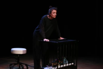 Une femme habille en noir sur une scène de théâtre avec les mains appuyées sur la table devant elle