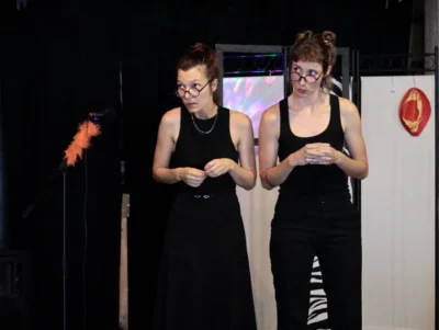 Sur une scène de théâtre, deux jeunes femmes avec des lunettes de vue, les bras dans la même position, et habillées en noir