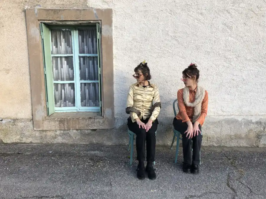 Deux jeunes femmes, assises sur des chaises dans la rue, regardent du côté droit, avec les mains sur les genoux