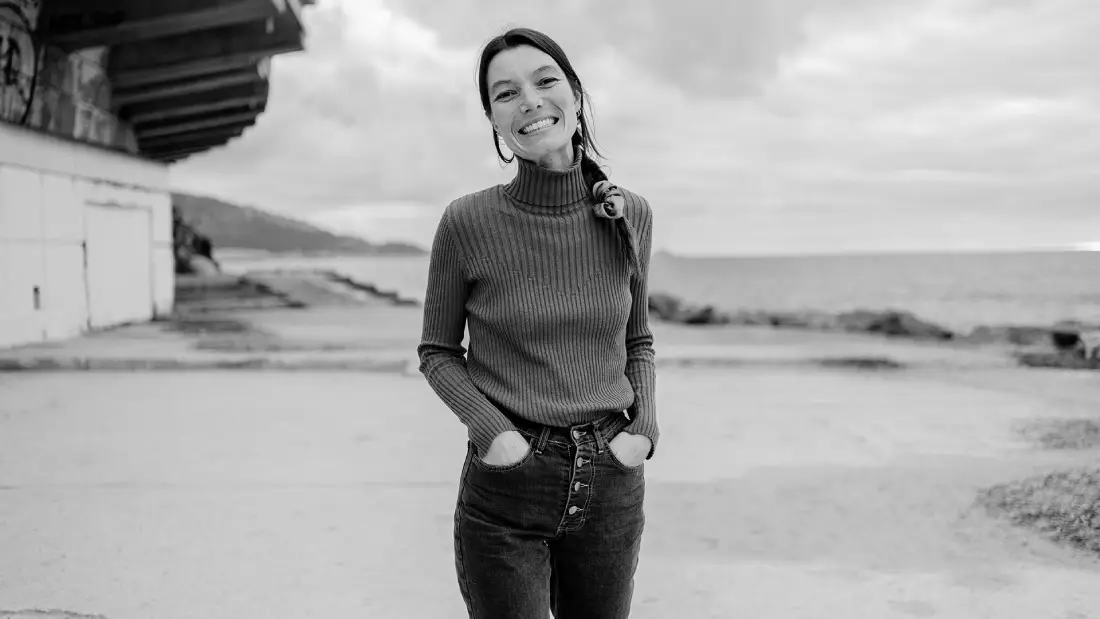Une jeune femme aux cheveux longs, souriante, en extérieur avec les mains dans les poches