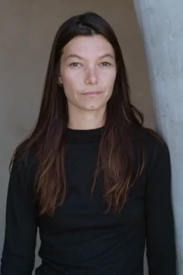 Une jeune femme avec un pull noir et les cheveux long à côté d'un mur blanc